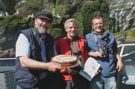 Dunter III skipper Jonathan Wills (left) presents Swiss tourist Andy Baumgartner with “The Birds of Shetland” book and cake to celebrate the 30,000th passenger with Seabirds-and-Seals, accompanied at the Noss gannetry with guide Jon Dunn (right) of Shetland Wildlife.