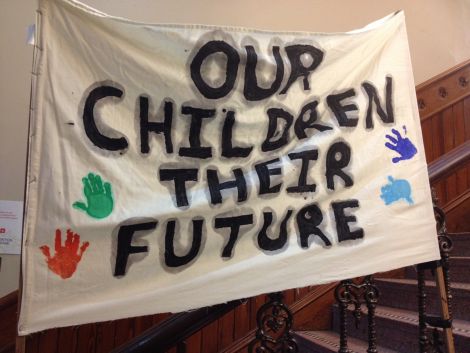 Protesters against school closures, listening to proceedings in the chamber on video link upstairs, have hung a banner on the stairs within Lerwick Town Hall.