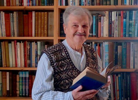 Elderly man with a mustache smiling while holding an open book, standing in front of a bookshelf filled with books.