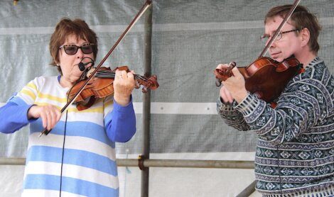 Two violinists, a woman in a striped shirt and a man in a patterned sweater, playing violins in an outdoor setting.