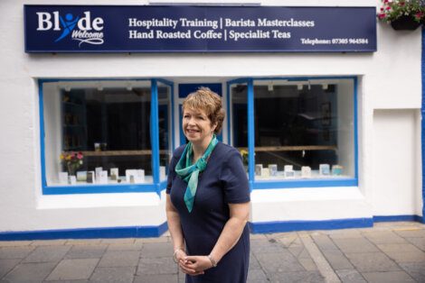 Woman standing in front of a coffee shop with a sign offering hospitality training and barista masterclasses.