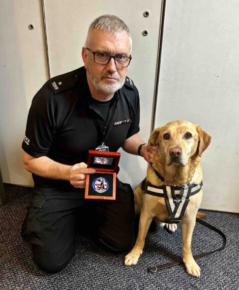 A man in a dark uniform kneels beside a labrador retriever, holding an award, both looking at the camera in a room with a metal door.