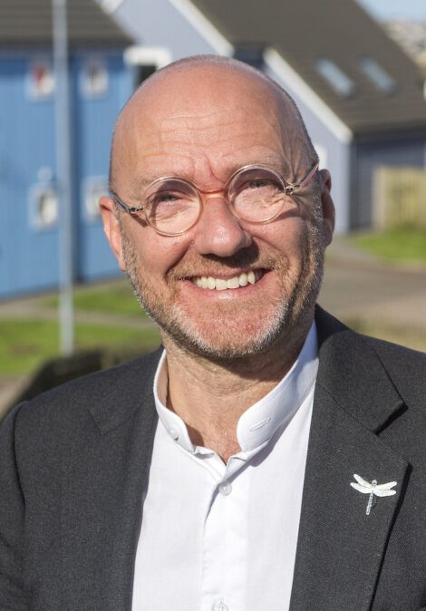 A smiling bald man with glasses wearing a suit jacket and white shirt, standing outside with houses in the background.