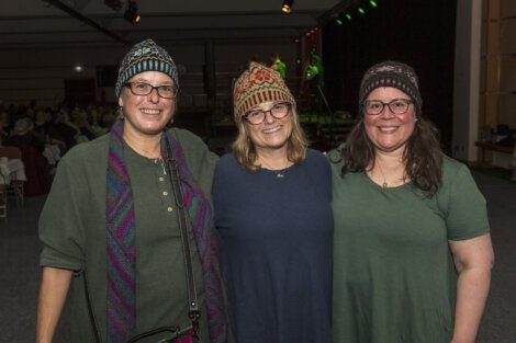 Three smiling women wearing knitted hats standing together at an event.
