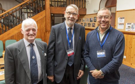 Three smiling men wearing conference badges posing in front of a staircase.