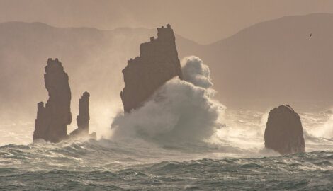Giant waves crashing against sea stacks in a misty, stormy ocean setting.