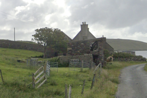 Rural stone house with a slanted roof near a paved road, with a wooden fence and green pastures under a cloudy sky.