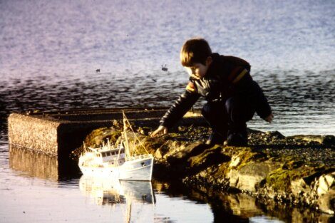 A boy playing with a toy boat by the water's edge.