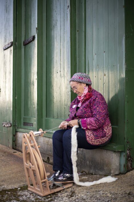 Elderly woman sitting by a green door, spinning wool with a traditional spinning wheel.