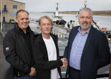 Three men shaking hands at a harbor with boats in the background.