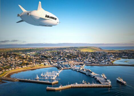 A white airship flying over a harbor and town.