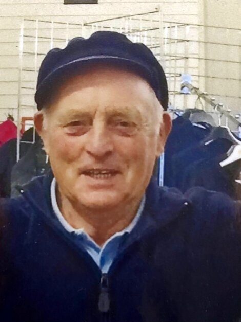 An older man in a hat is smiling in front of a rack of clothes.