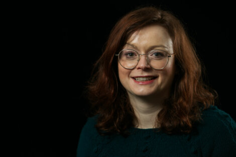 A woman with red hair and glasses is smiling in front of a black background.