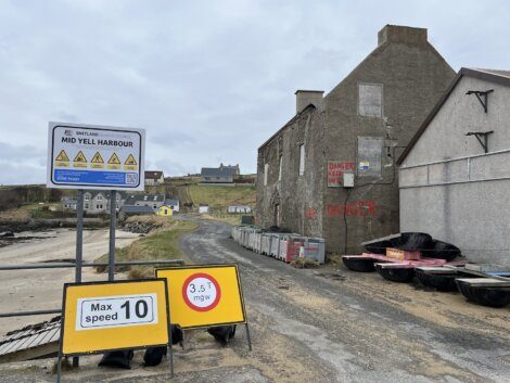 Road leading to mid yell harbour with speed limit signs and a dilapidated building with 'danger keep out' warnings.