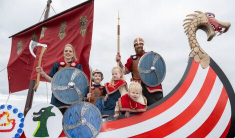 A family dressed as vikings with shields and a spear aboard a boat float with a dragon head design during a parade.