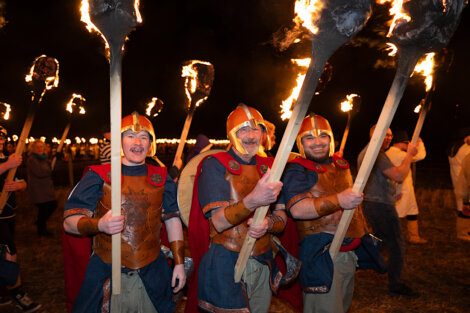 Participants in historical costumes holding flaming torches at a nighttime event.