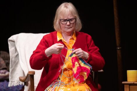 Woman knitting with colorful yarn while seated onstage.