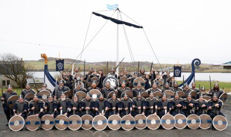 A group of people dressed as viking warriors with shields and helmets posing in front of a viking ship structure.