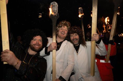 Three individuals dressed in wigs and white lab coats smiling at the camera, holding lit torches at a night event.
