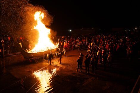 Crowd gathered at night around a large bonfire with a boat engulfed in flames.
