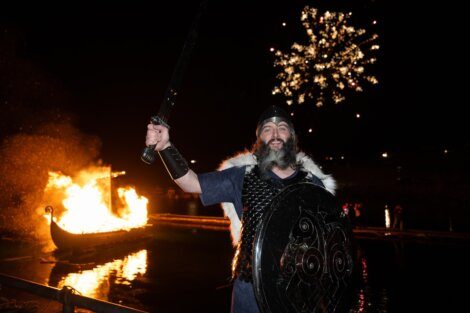 Man in viking costume holding a sword and shield with a burning boat and fireworks in the background.