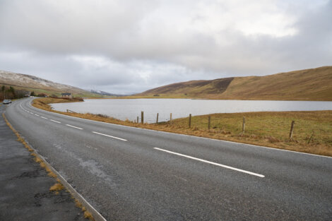 Curving road alongside a calm lake with gentle hills under a cloudy sky.