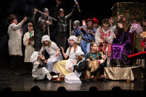 Children dressed in various costumes perform on stage in a theatrical production.