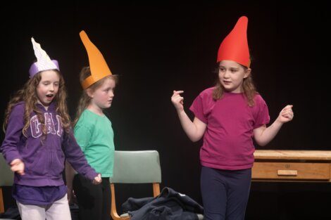Three children performing on stage, with one wearing a whimsical pointed hat.