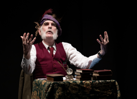 A person performing on stage, dressed in vintage attire with books and a pocket watch on a table, portraying a dramatic expression with raised hands.