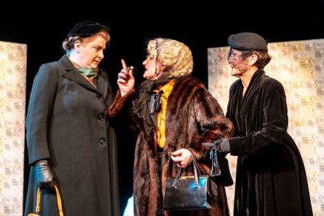 Three actors on stage portraying an intense conversation, with one actor gesturing emphatically.