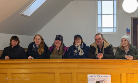 A group of seven adults smiling behind a wooden railing indoors.