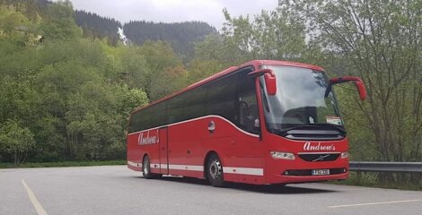 A red andersson tour bus parked on a roadside with green trees and a hill in the background.