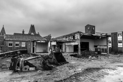 Black and white photo of a dilapidated building with a collapsed roof and scattered debris, featuring a toppled excavator in the foreground.