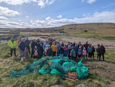 A large group of people of various ages posing for a photo during a community clean-up event, with a pile of collected trash and nets in the foreground and a rural landscape in the background.