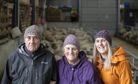 Three generations of a family—a senior man, senior woman, and younger woman—smiling in a barn with sheep in the background.