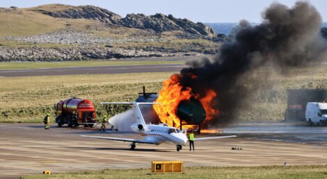 Aircraft emergency training simulation at an airport, featuring a jet with controlled fire near its engine as fire personnel oversee the exercise.