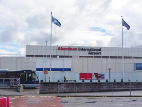 Front view of aberdeen international airport building with two scottish flags flying and a shuttle bus outside.