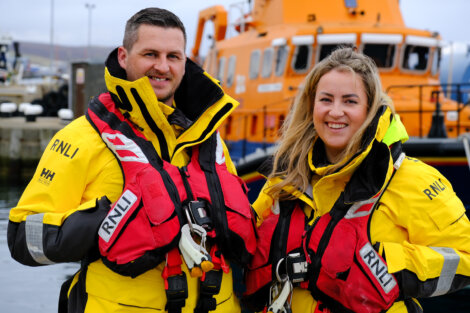 Two lifeboat crew members in yellow rnli jackets smiling in front of a lifeboat.